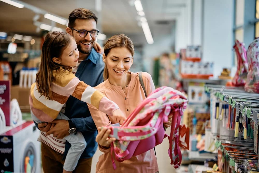 Anne, baba ve kız çocuğu kırtasiye mağazasında birlikte okul çantası seçiyor — okul alışverişinde dikkat edilmesi gerekenler