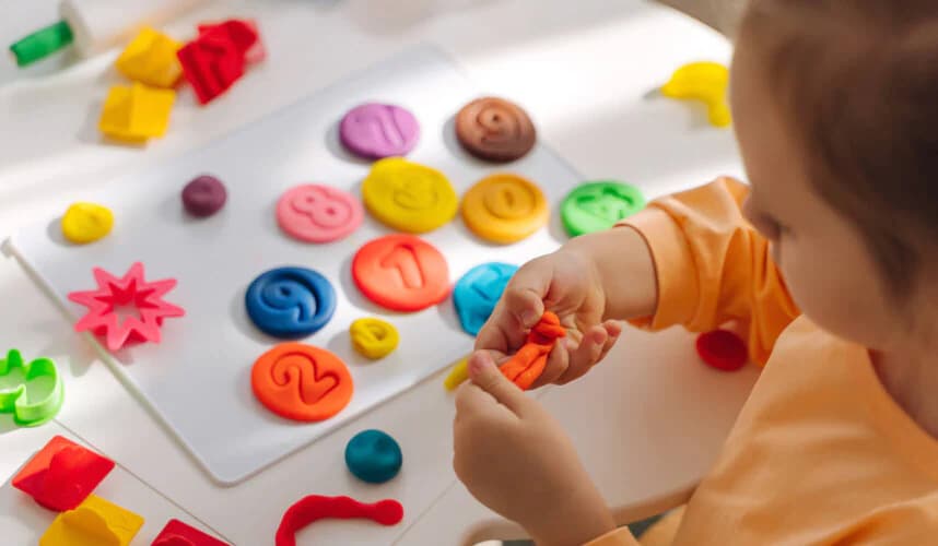 Masa başında oyun hamuruyla renkli sayılar ve şekiller yapan, okul öncesi yaşta bir çocuk.