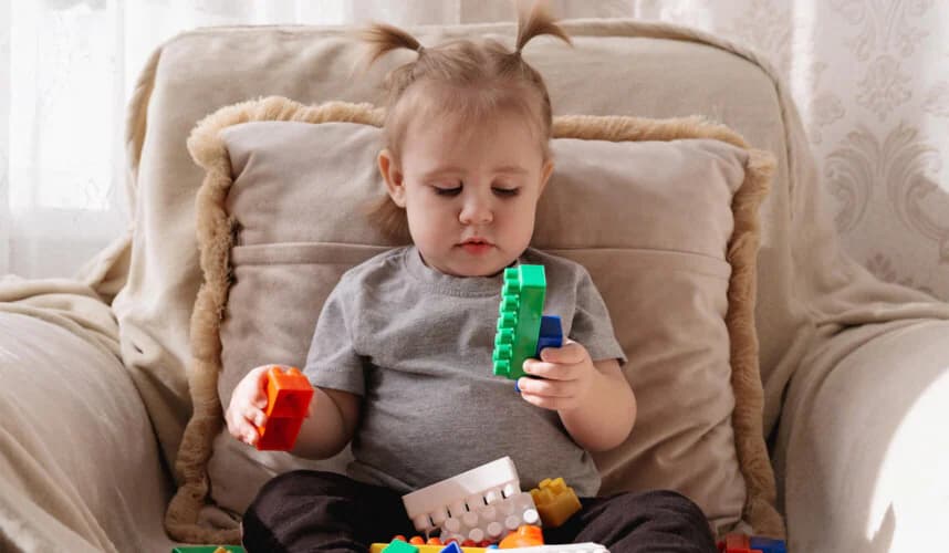 Koltukta oturan küçük çocuk, renkli yapı bloklarıyla odaklanarak oynuyor; oyunla öğrenme ve bilişsel gelişim aktivitesi.