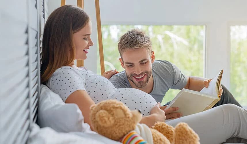 Hamile bir kadın ve baba adayı, evde yatağın üzerinde oturup karnındaki bebeğe kitap okurken gülümsüyor; önde pelüş ayı görülüyor