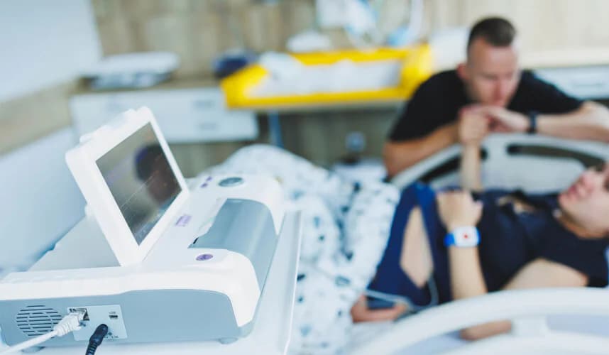 Doğum odasında fetal kalp atım monitörü önde net görünürken, arka planda yatağında yatan anneye elini tutarak destek olan baba