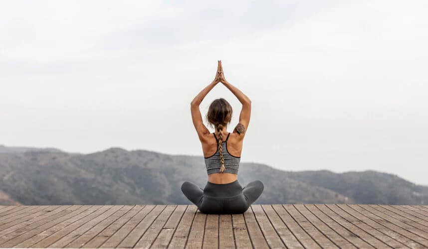 Dağ manzarasına karşı ahşap terasta bağdaş kurup ellerini baş üstünde birleştiren kadın; açık havada yoga ve meditasyon pratiği