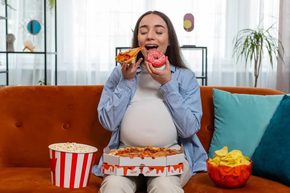 hamilelikte aşerme yaşayan hamile bir kadının pizza ve tatlı yiyeceklerle koltukta oturduğu görsel